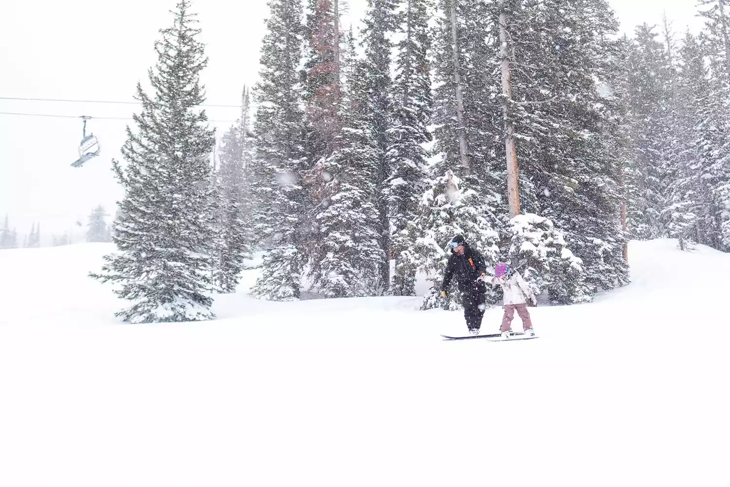 An adult and child snowboarding side-by-side at Brighton Resort in Utah during a sunny day.