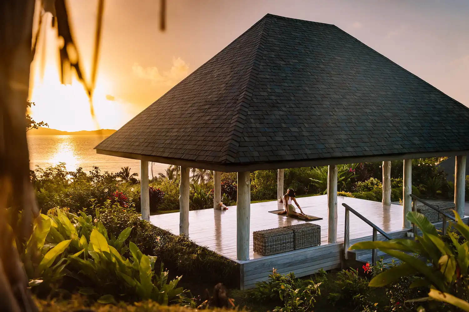 A person practicing yoga at sunset in an open-air wooden pavilion overlooking the ocean.