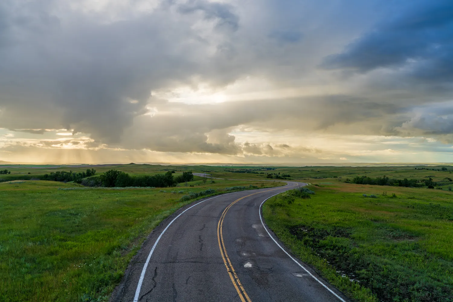 A smooth, winding asphalt road cutting through vast golden grasslands under a bright, cloud-streaked sky.