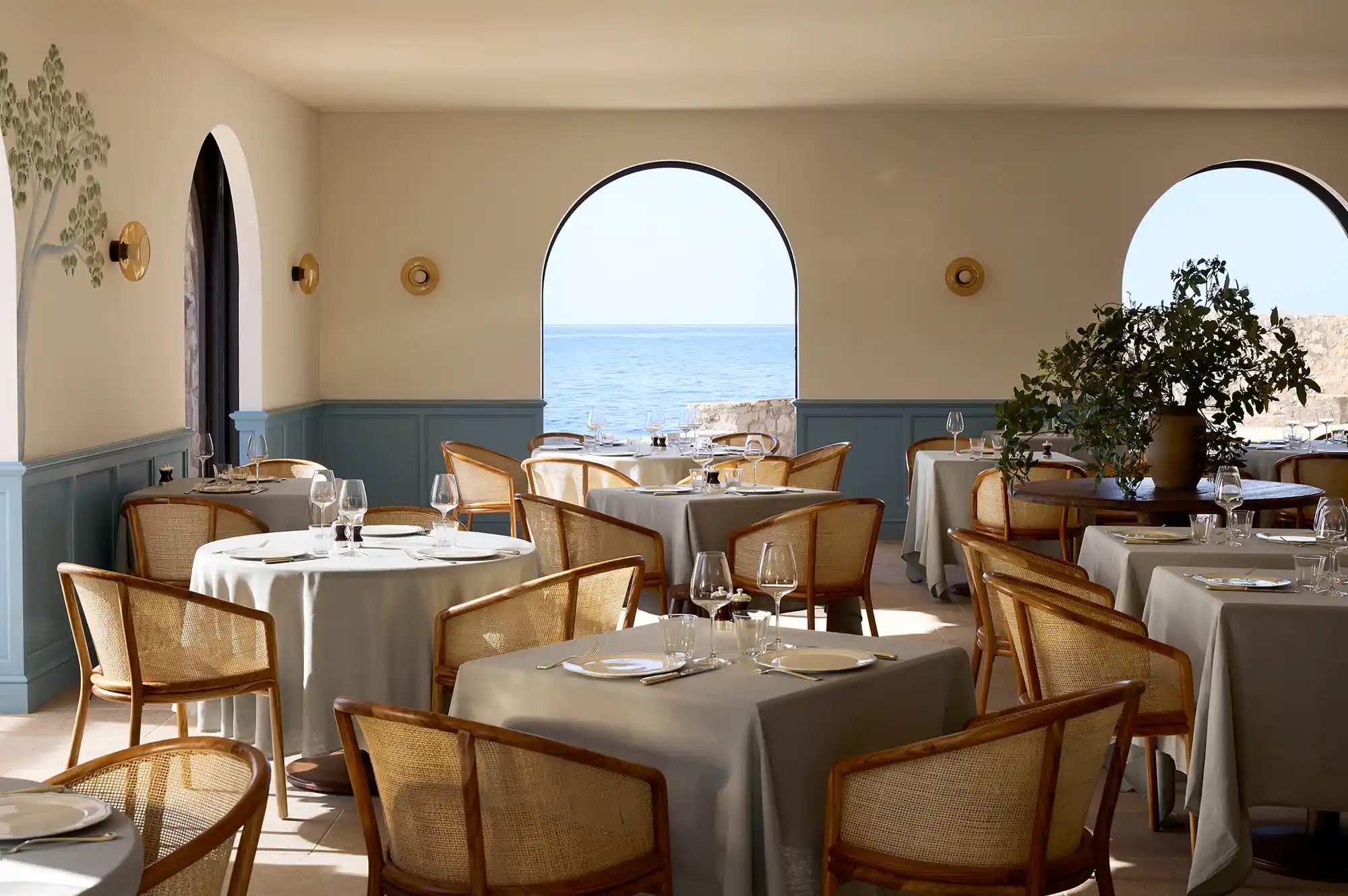 Upmarket dining room with wicker chairs and large arched windows overlooking the sea.