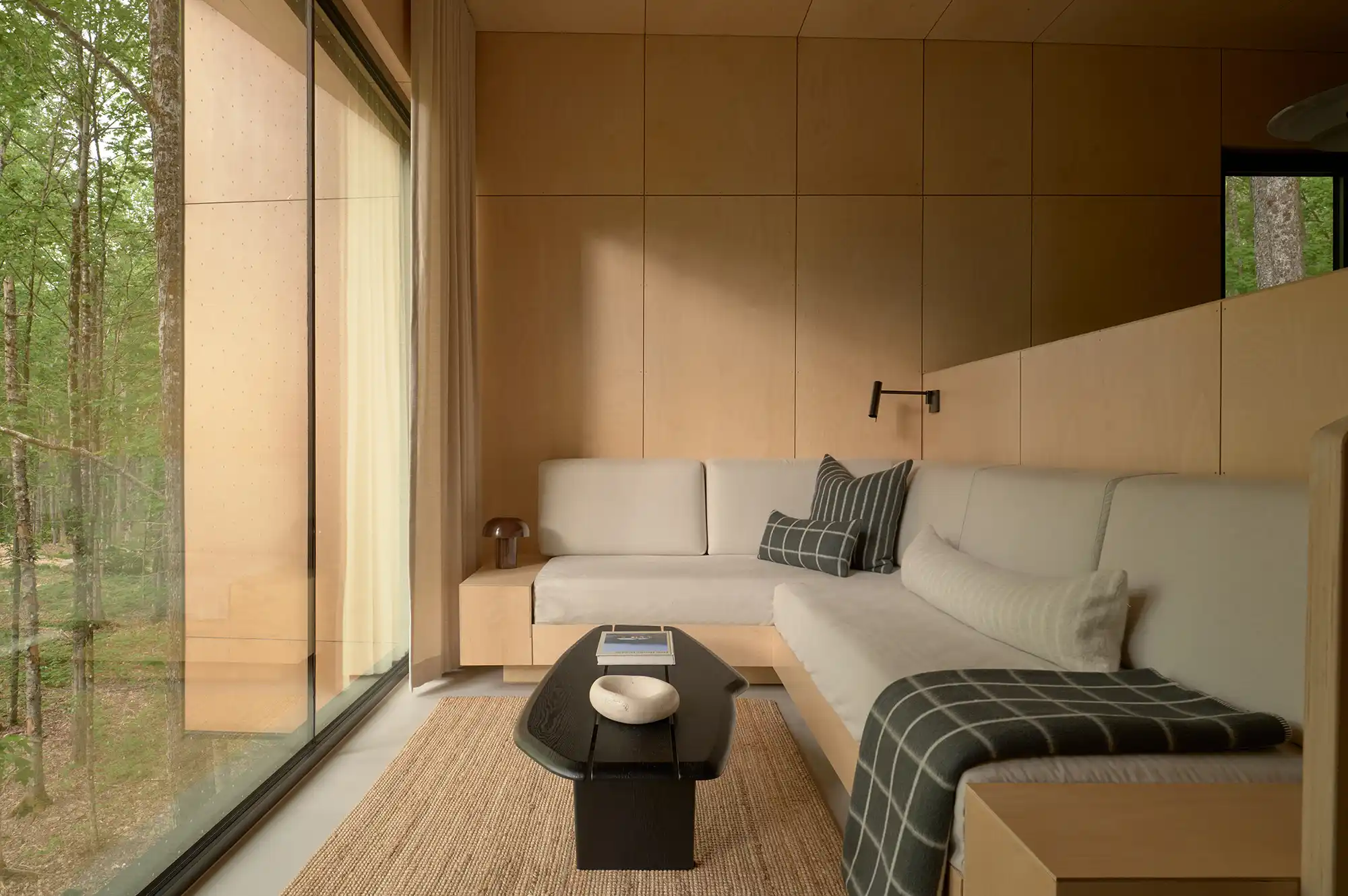 Interior of a modern living room with light wood walls, a grey sectional sofa, and a large window looking out to trees.