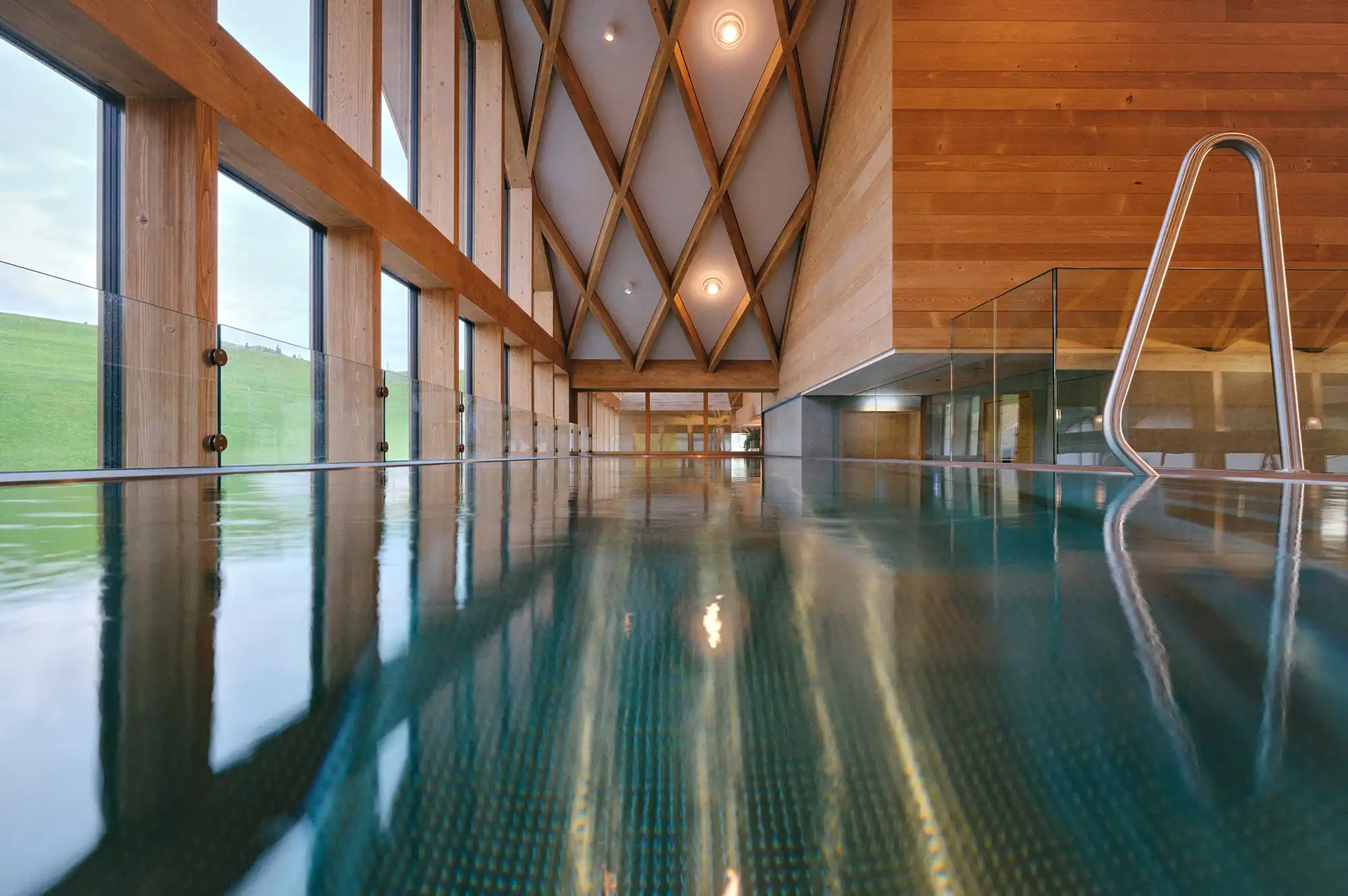 Indoor infinity pool surrounded by light wood panels with large windows overlooking the rolling green hills of Appenzell.