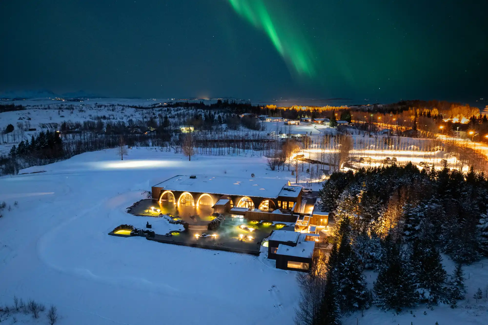 Aerial view of Laugarás Lagoon at night under the Green Northern Lights with snow on the ground.