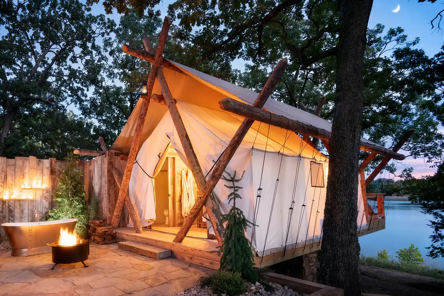 A glamping tent at Camp Long Creek featuring an outdoor soaking tub and fire pit under a canopy of trees.