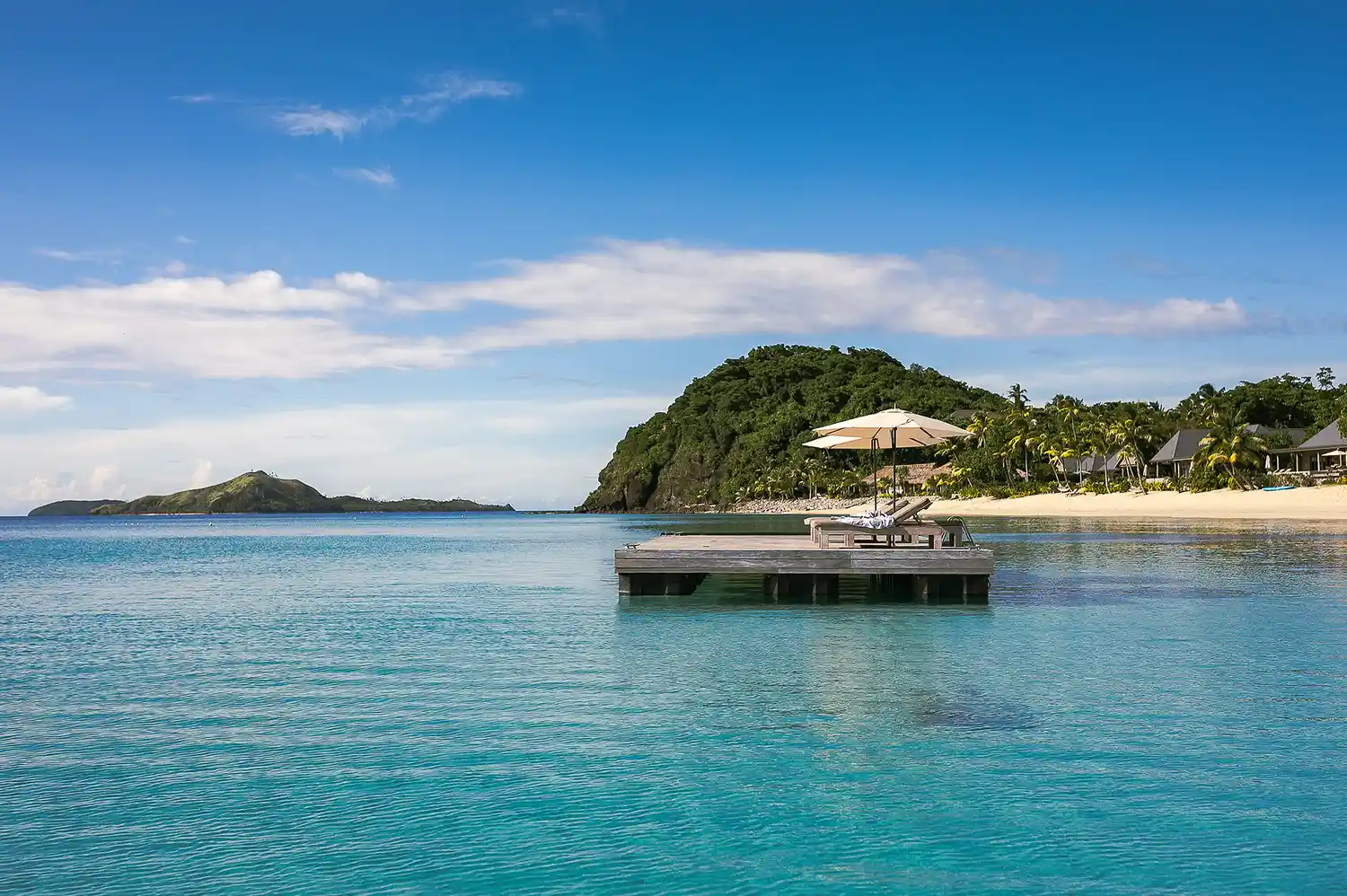 A wooden floating pontoon with white lounge chairs overlooking the crystal clear ocean and the tropical island of Kokomo.