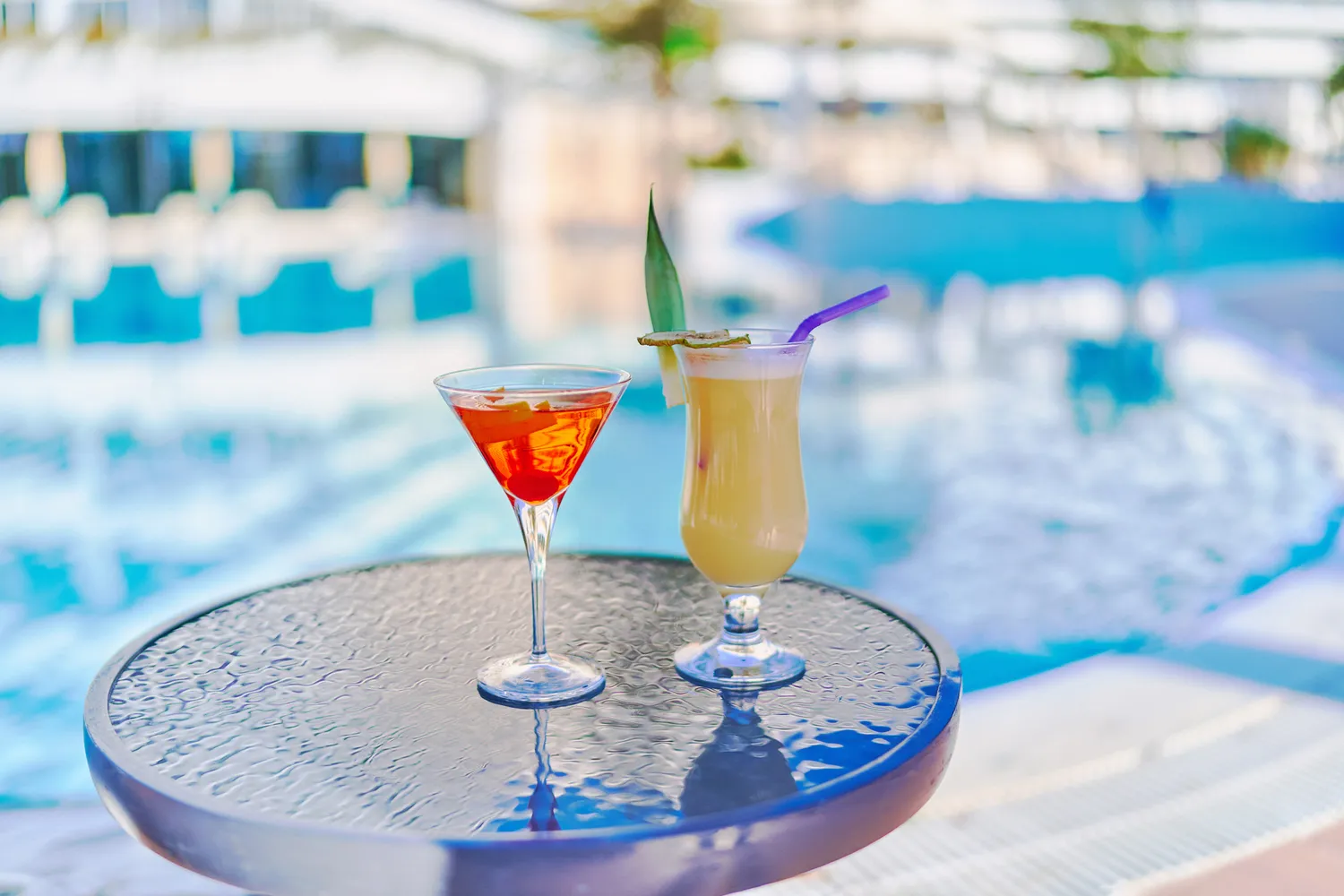 Two colorful tropical cocktails sitting on a table by a resort swimming pool.