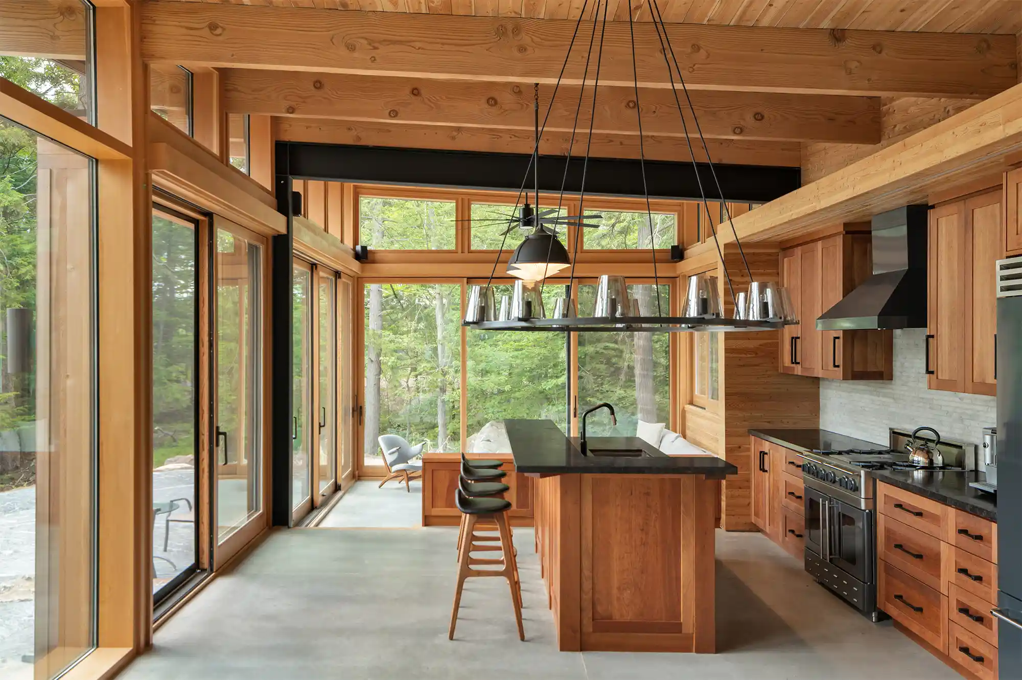 Modern kitchen with light wood cabinets and a central island with views of the forest through large windows.