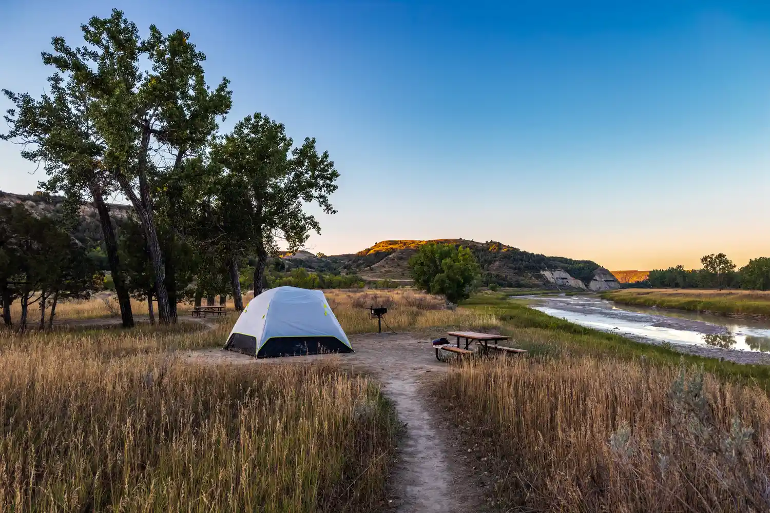 A blue tent pitched at a campsite with a picnic table and grill near a tree-lined riverbank.