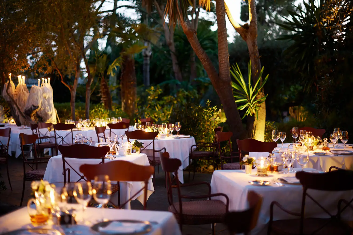 An elegant outdoor restaurant setting under trees with warm ambient lighting at night.