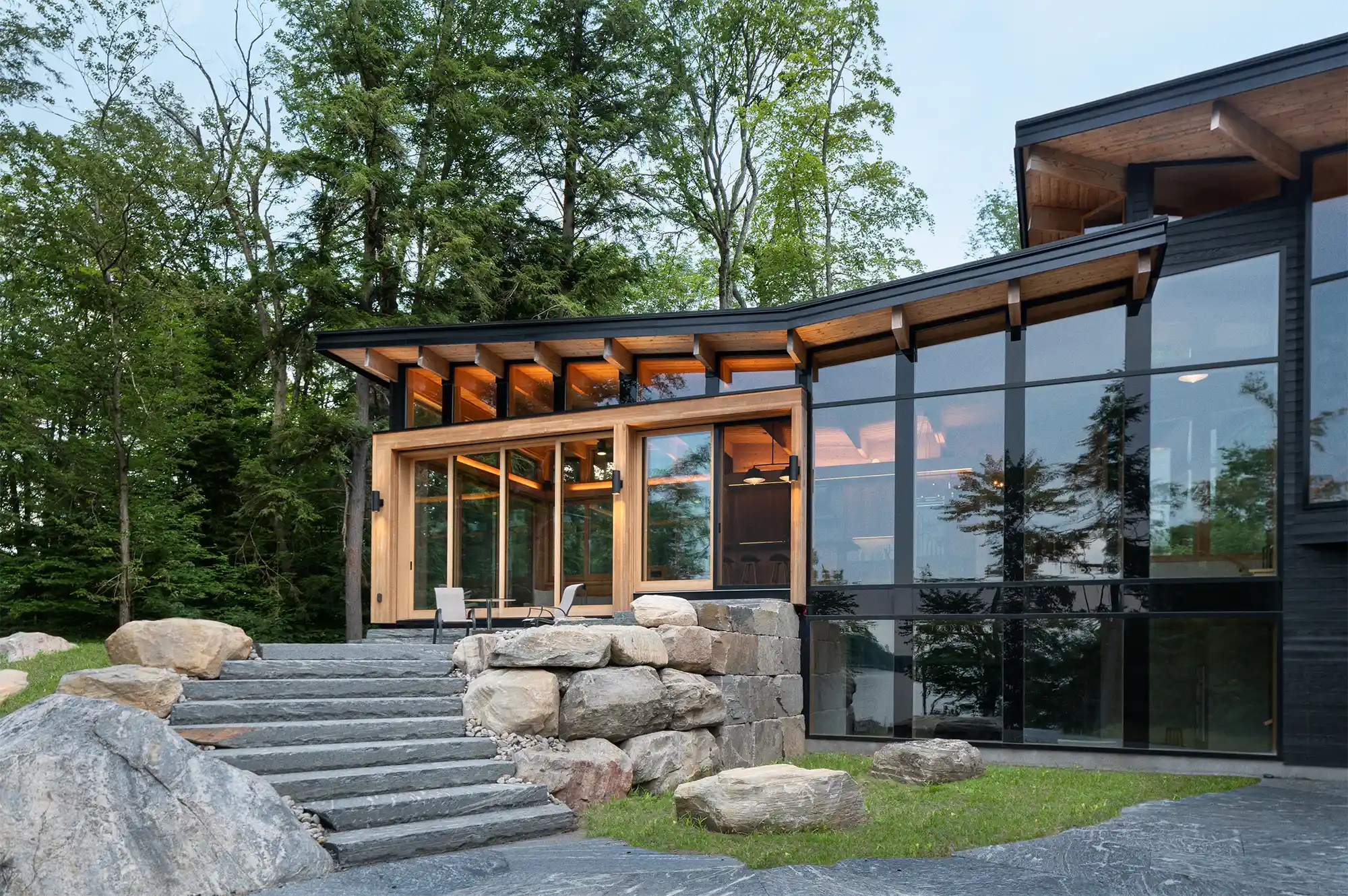 Exterior of a modern cottage with a glass facade and stone steps surrounded by dense forest.