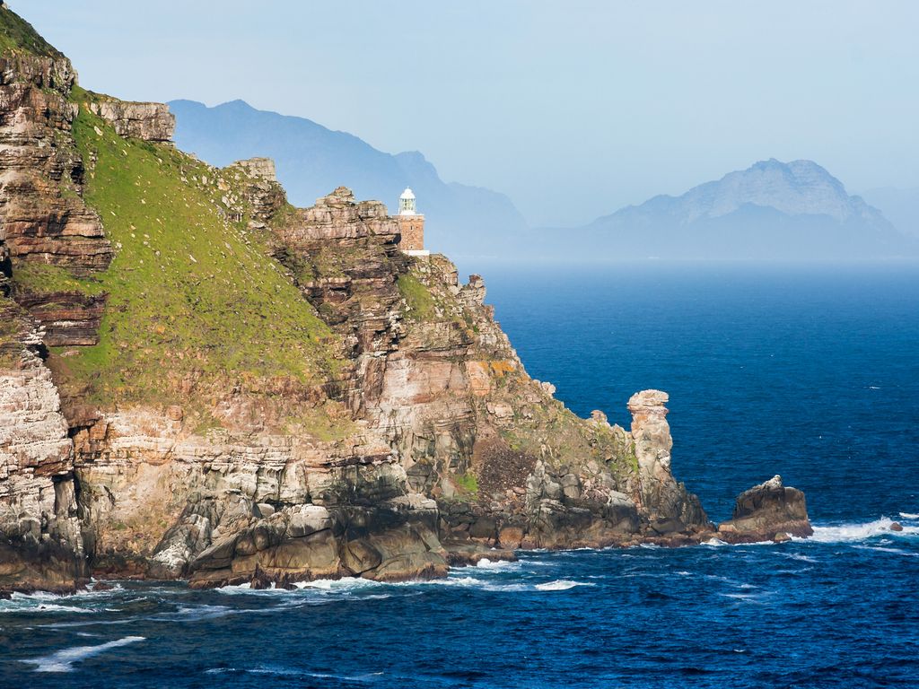 A lone lighthouse perched on a steep cliff overlooking the turbulent Atlantic Ocean at Cape Point.
