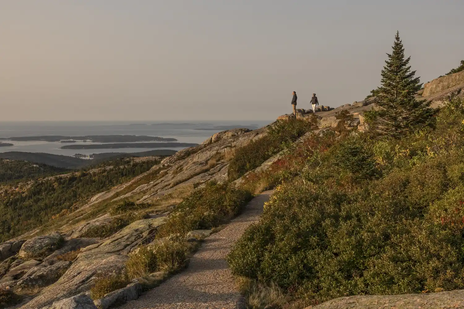 A paved mountain summit pathway with people looking out over a scenic bay filled with islands.