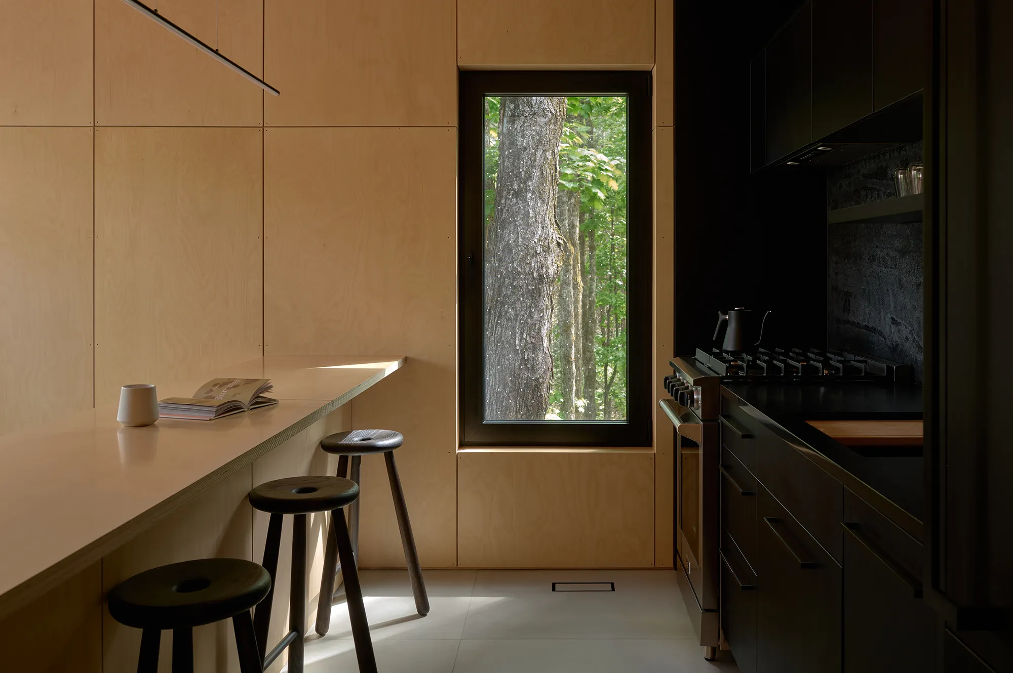 A modern kitchen with wood grain cabinetry, a large window view of the forest, and bar seating.