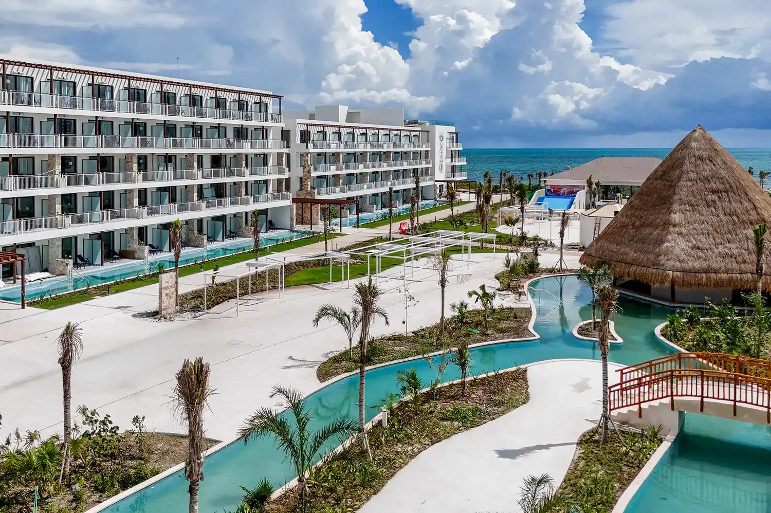 Contemporary tropical resort exterior featuring a large pool area and thatched-roof lounge near the ocean.