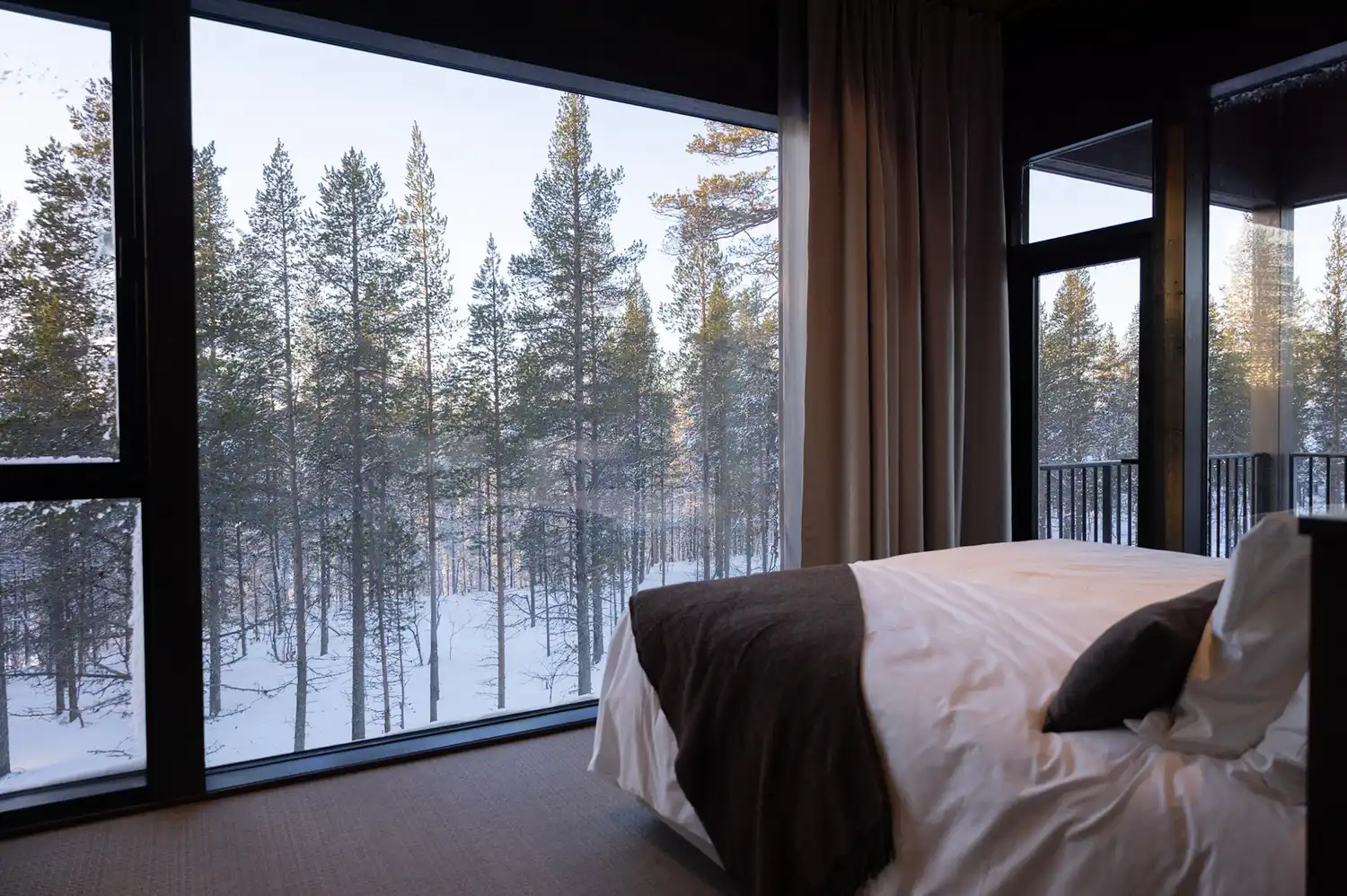 A luxury hotel suite featuring floor-to-ceiling glass walls overlooking a snowy evergreen forest.