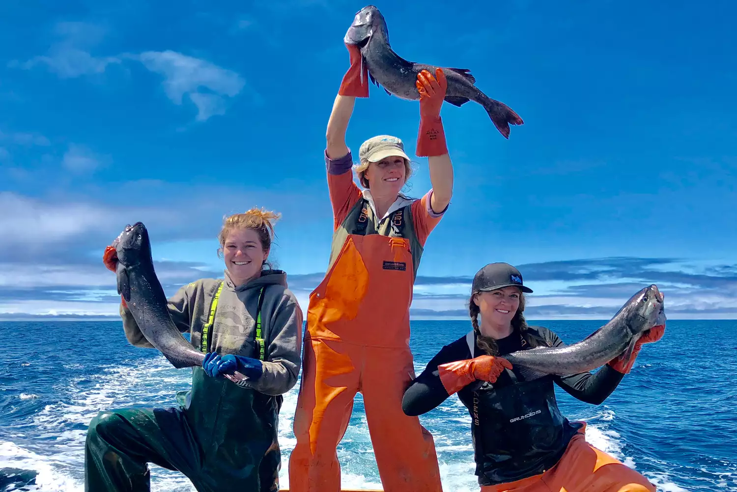 Three women smiling while holding up large, fresh fish at the Princess Seafood market in Noyo Harbor.