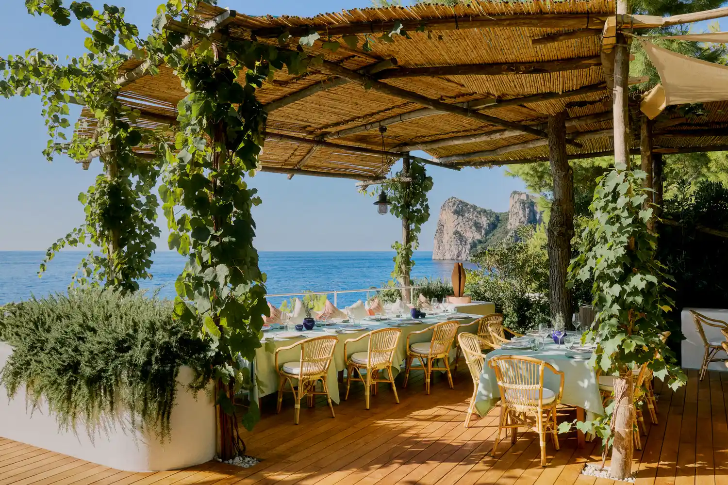 Outdoor restaurant terrace with tables under a vine-covered pergola overlooking the turquoise sea and distant cliffs.