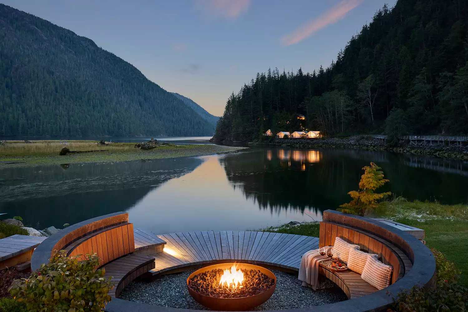 Adirondack chairs gathered around a flickering stone fire pit at the water's edge.