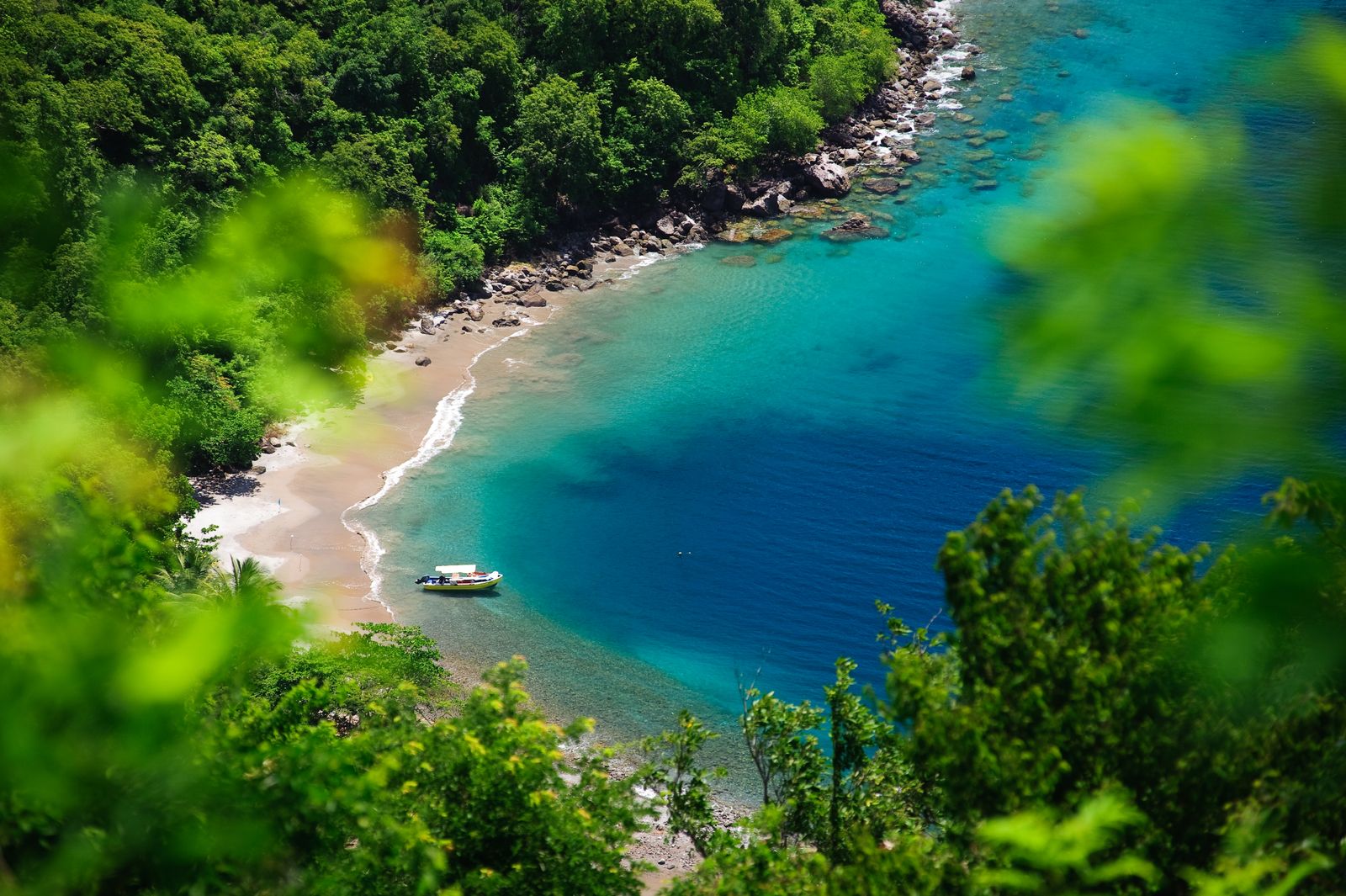 An aerial view of a boat floating on crystal clear turquoise Caribbean water near a white sand shoreline.