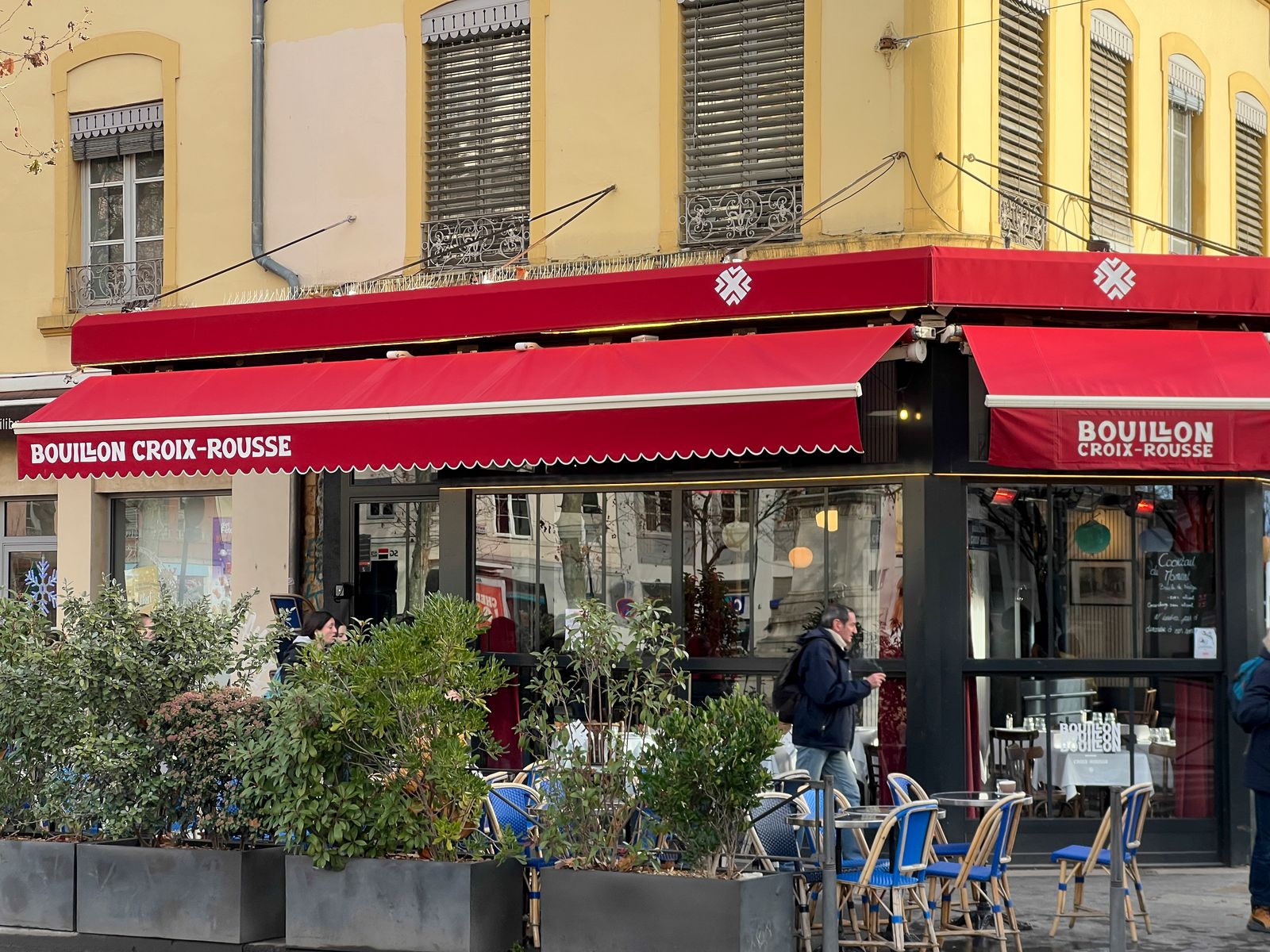 People walking past a street-side cafe with awnings in a Lyon neighborhood.
