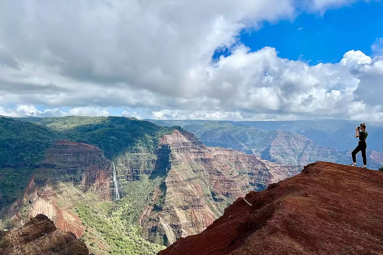 Hikers on a trail overlooking the red dirt and deep green ridges of Waimea Canyon.