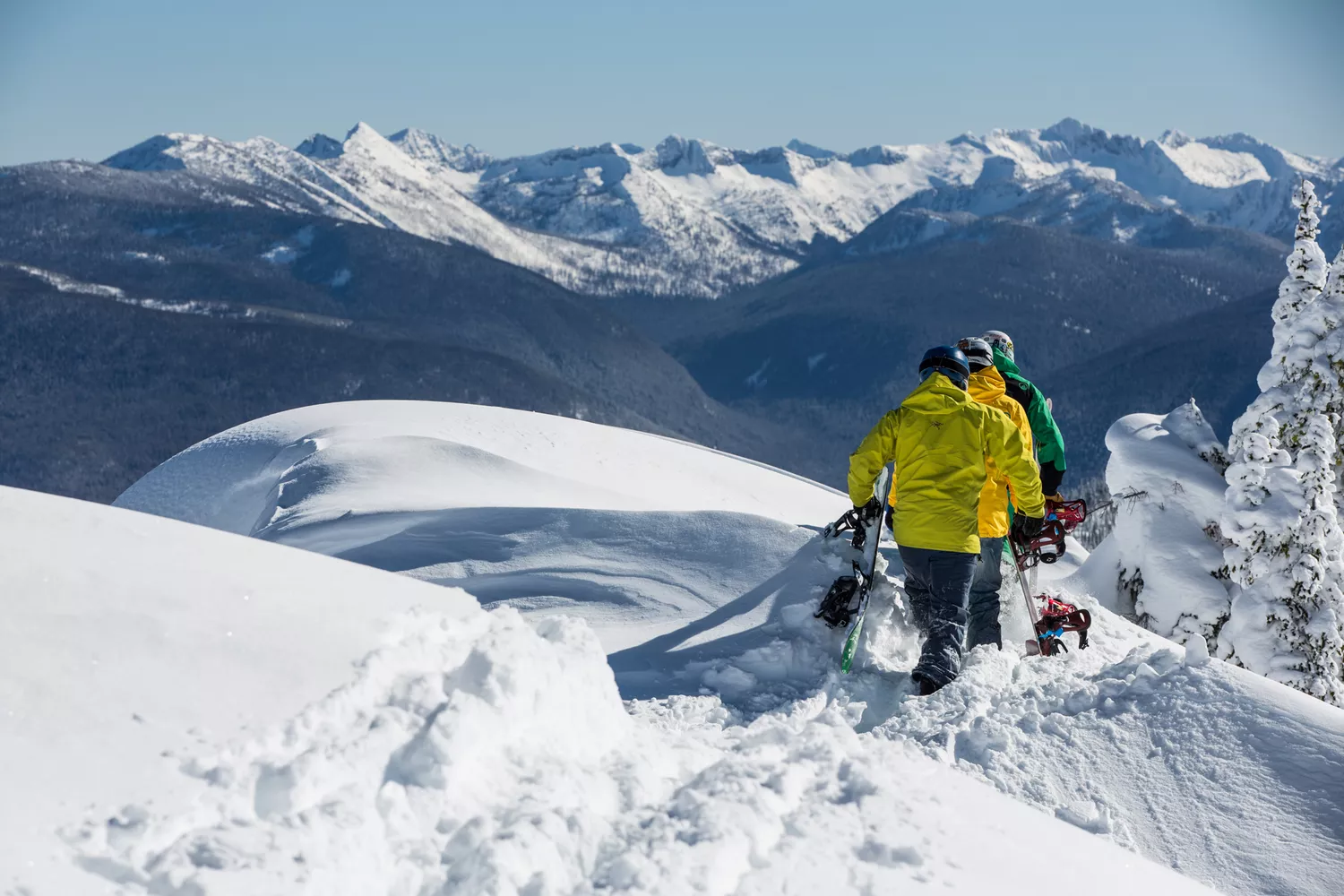 A group of snowboarders carrying their boards while hiking along a snowy mountain ridge in British Columbia.