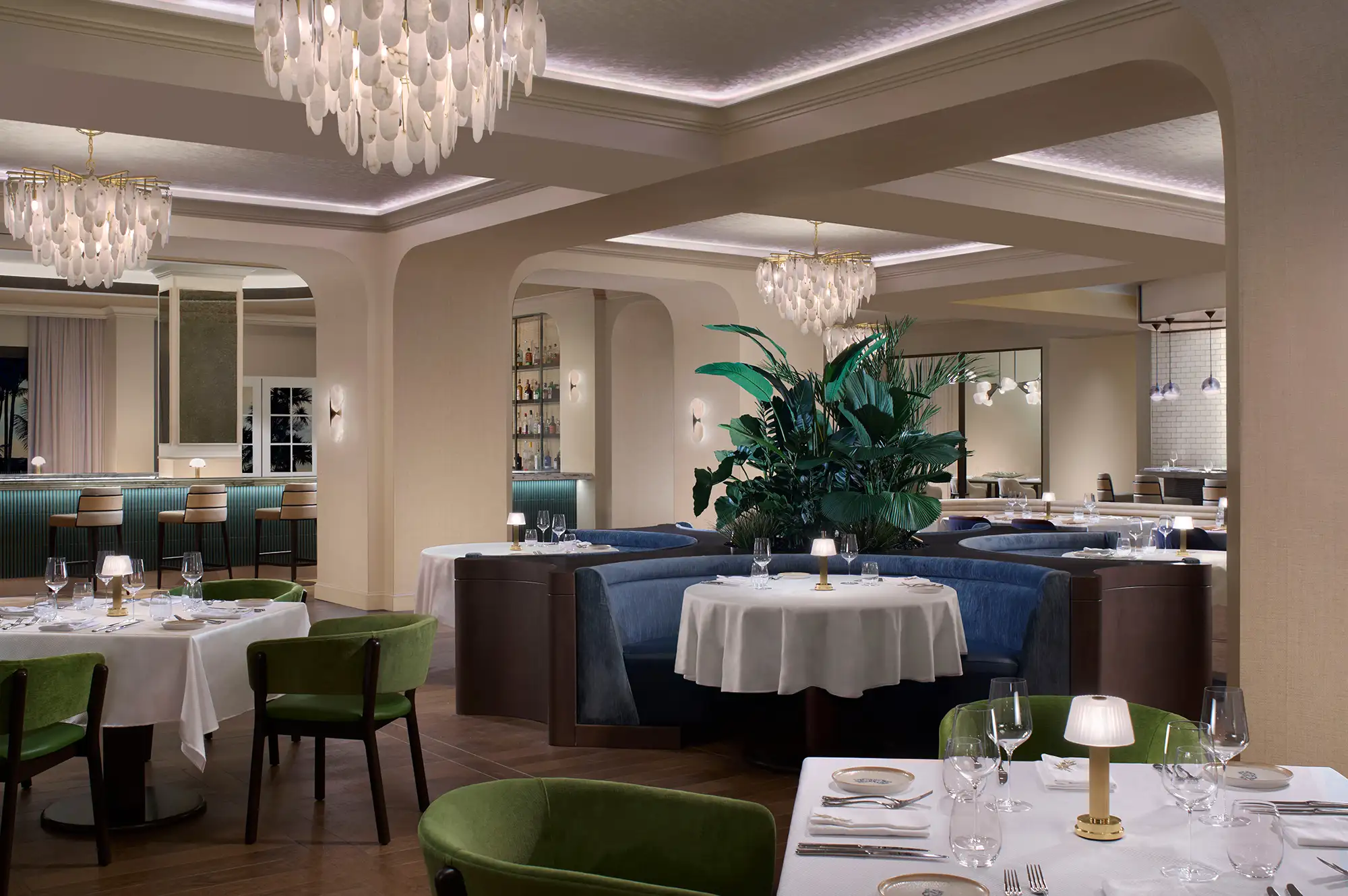 Upscale dining room featuring green velvet chairs, chandeliers, and a central indoor plant arrangement.