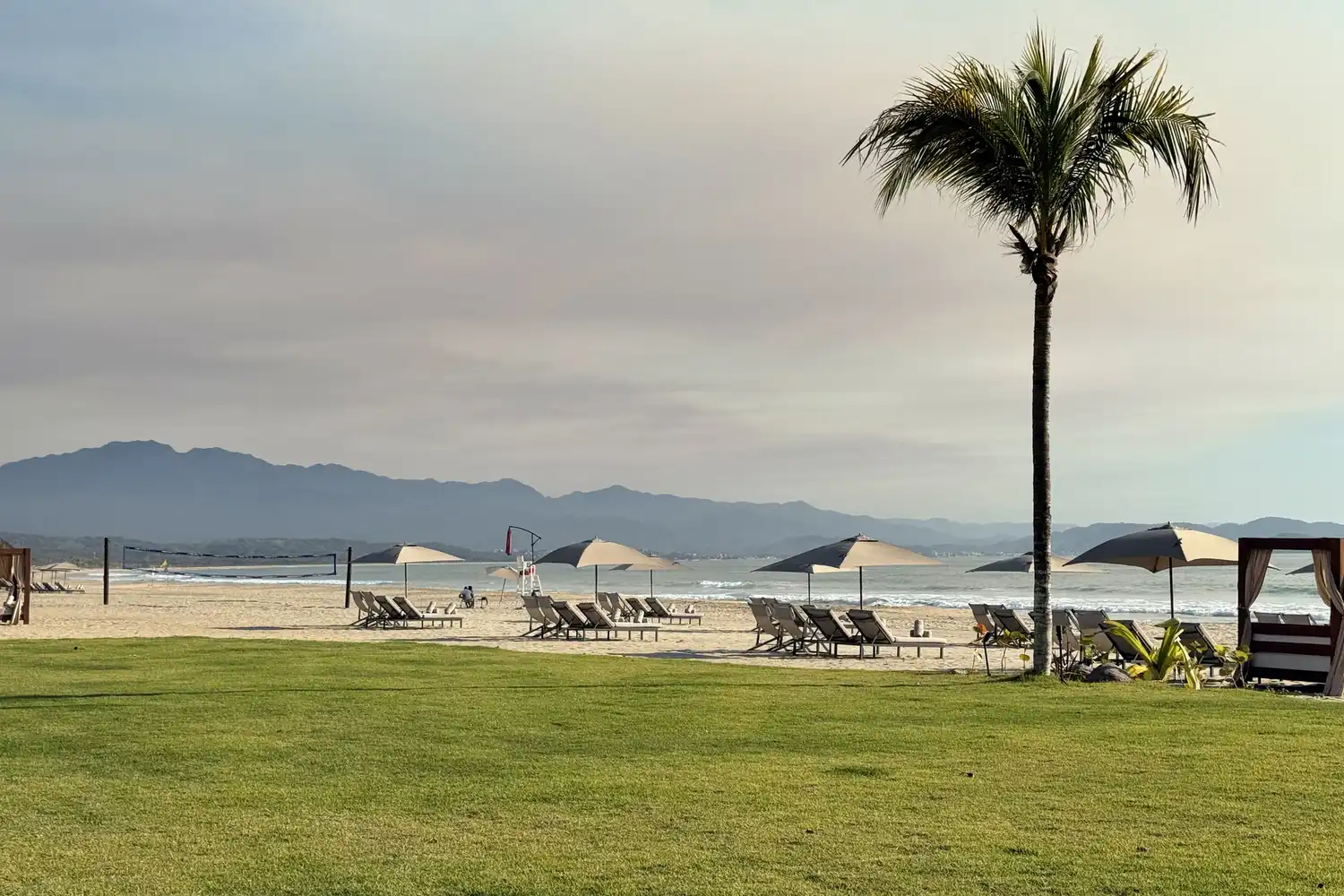 A wide golden sand beach with lounge chairs, ocean waves, and palm trees.