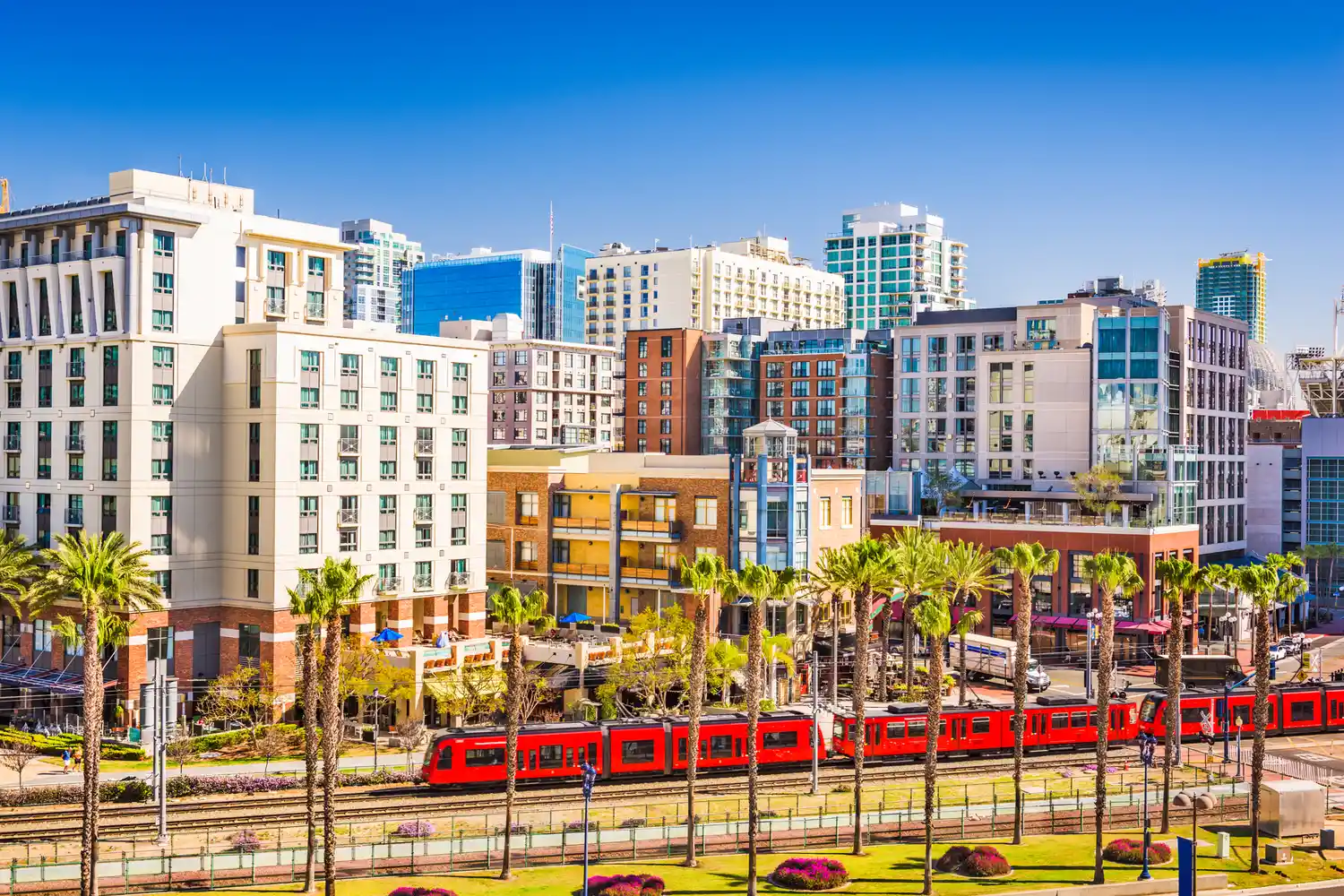 A bright red trolley car traveling through a downtown San Diego street with skyscrapers in the background.