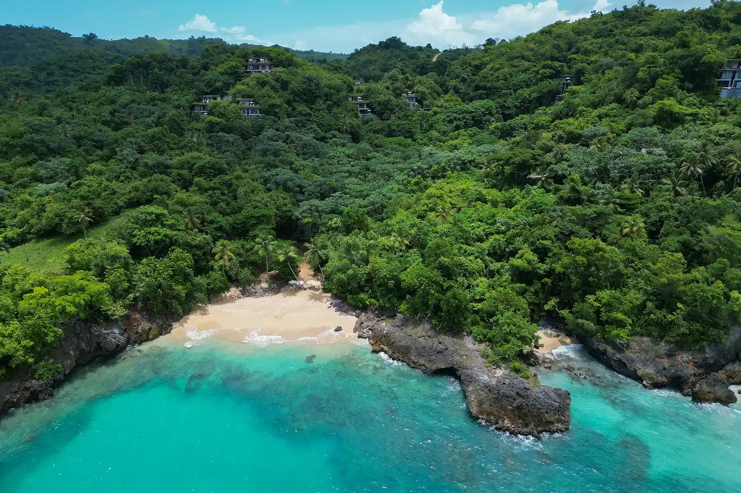 Aerial overview of a lush tropical beach with turquoise water and forested hills.