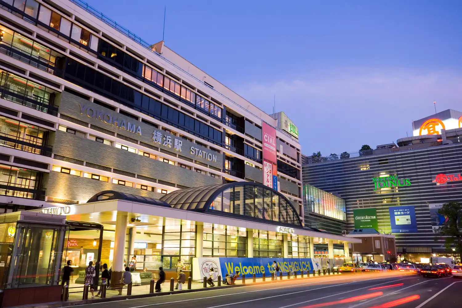 The modern exterior and west exit of Yokohama Station with lit signs.