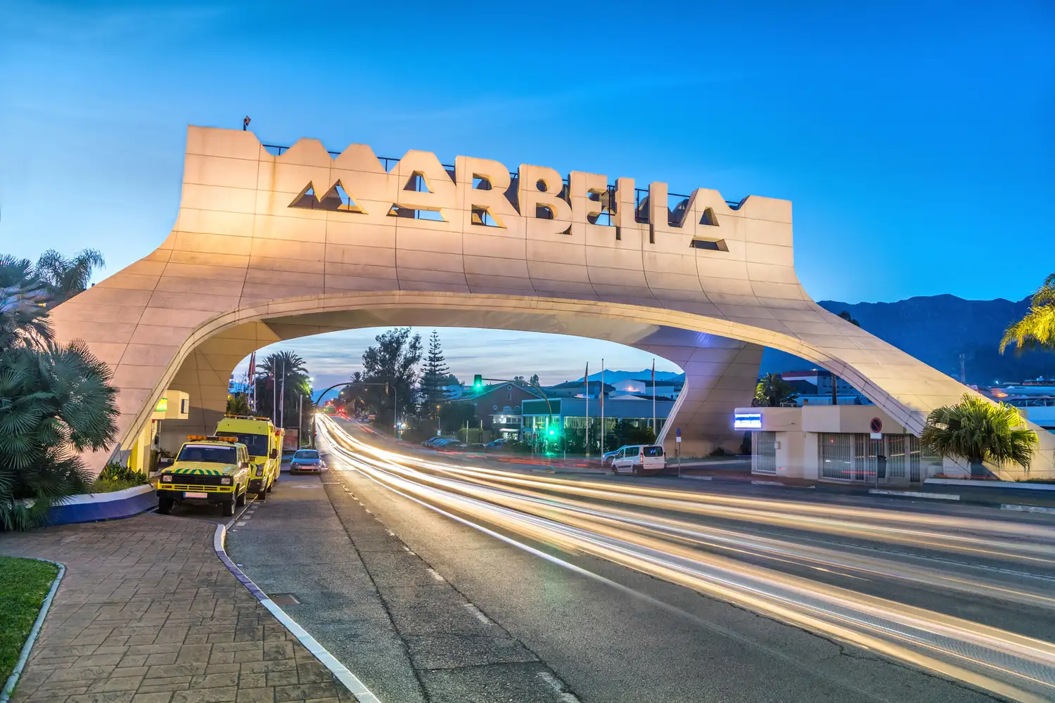 The iconic Marbella entrance arch illuminated at dusk with vehicle light trails.
