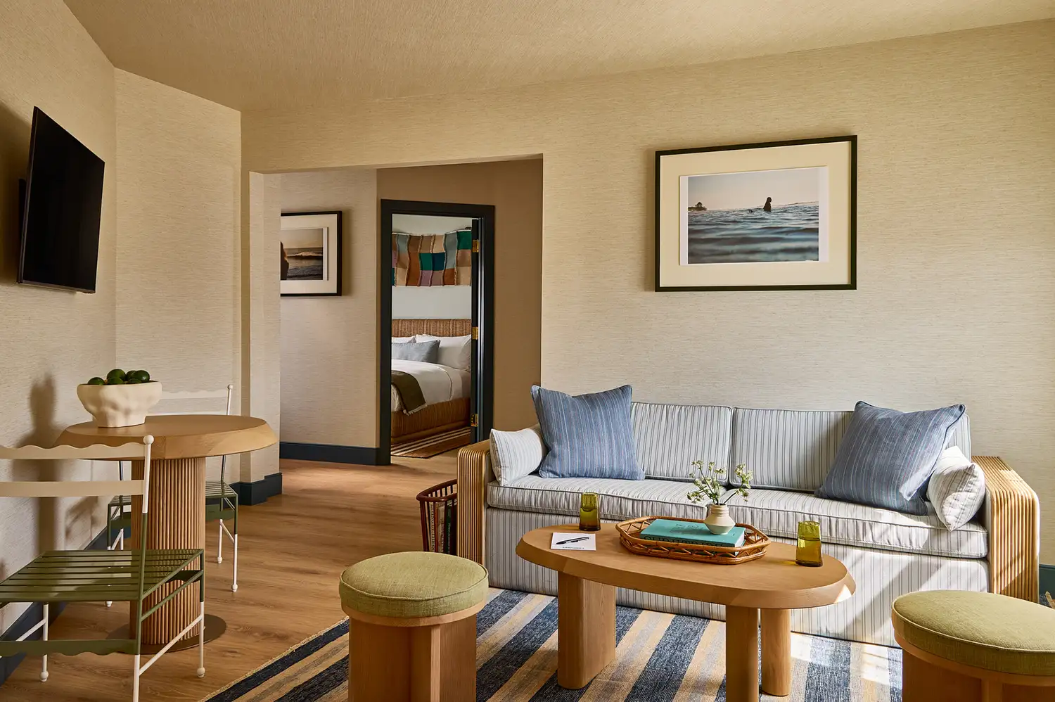 A hotel suite living room with a neutral sofa, warm wooden furnishings, and a view into the adjoining bedroom.