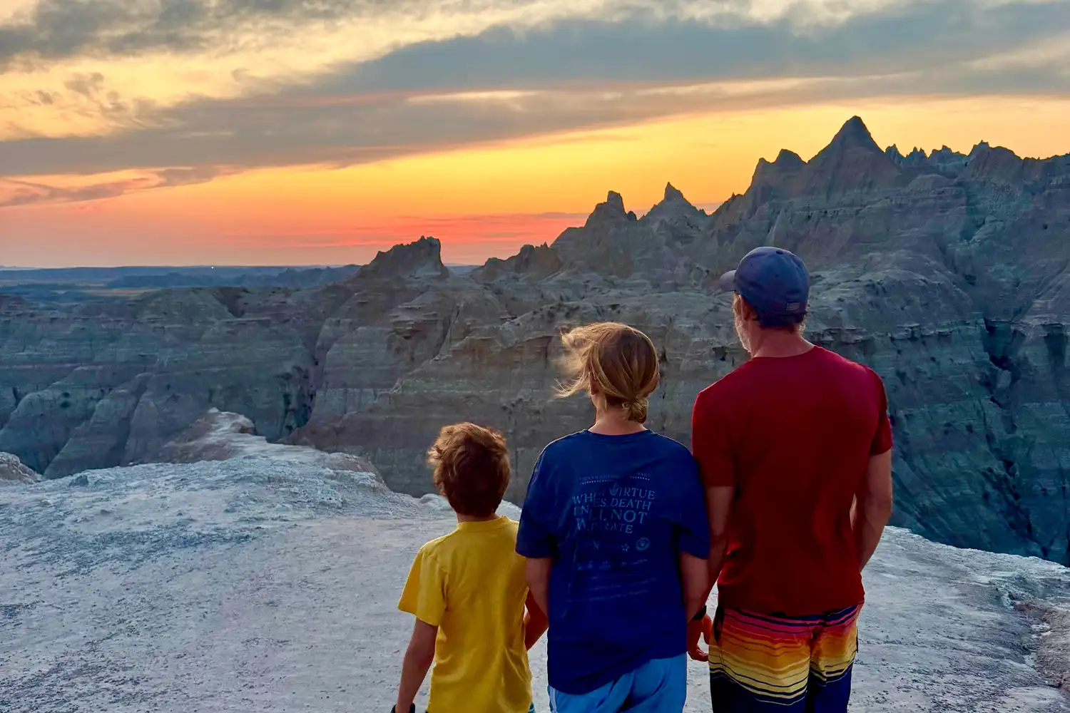 A family of three standing on a rocky ledge watching the sunset over a hilly park landscape.