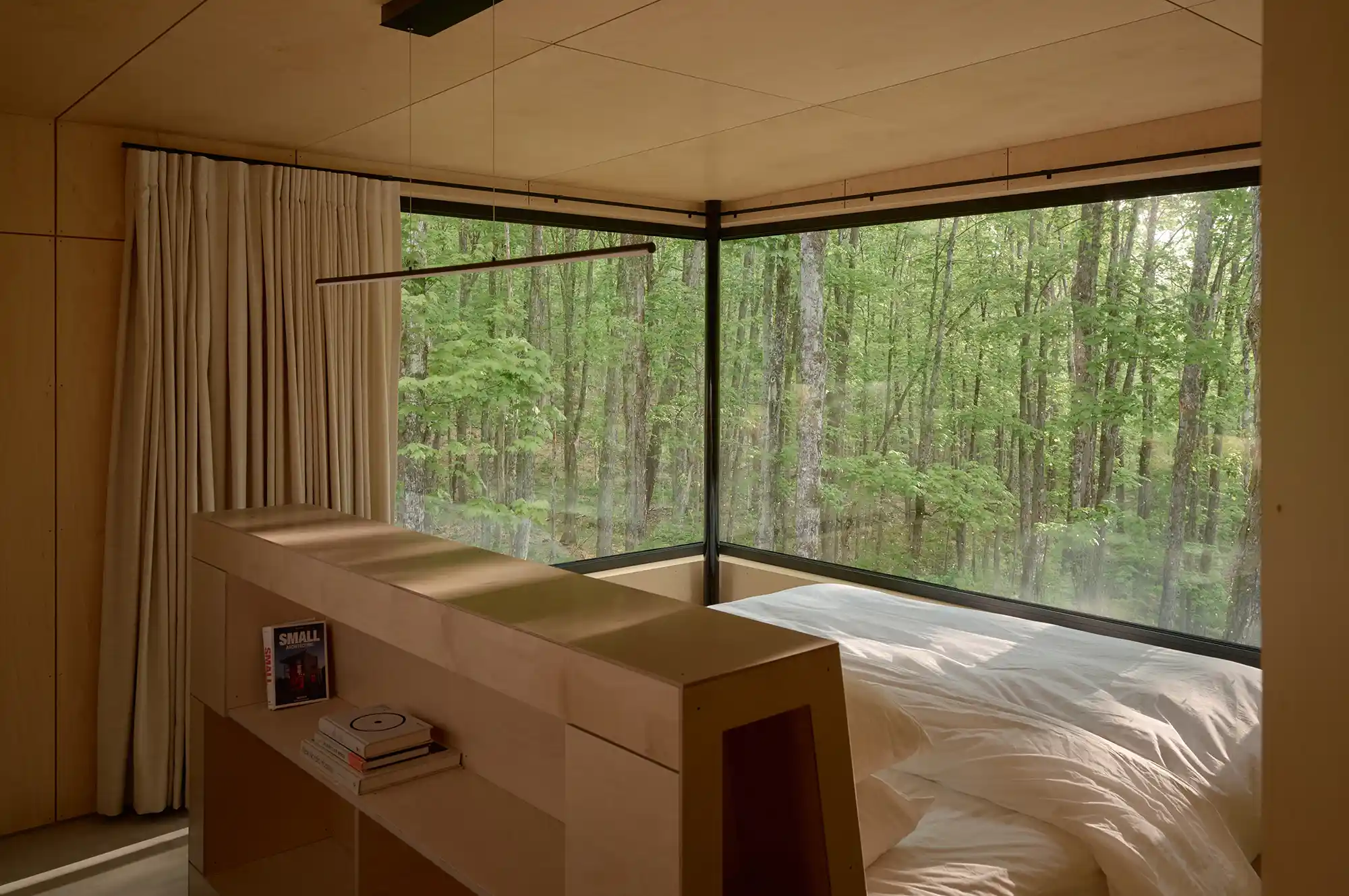A minimalist bedroom featuring floor-to-ceiling corner windows overlooking a lush deciduous forest.