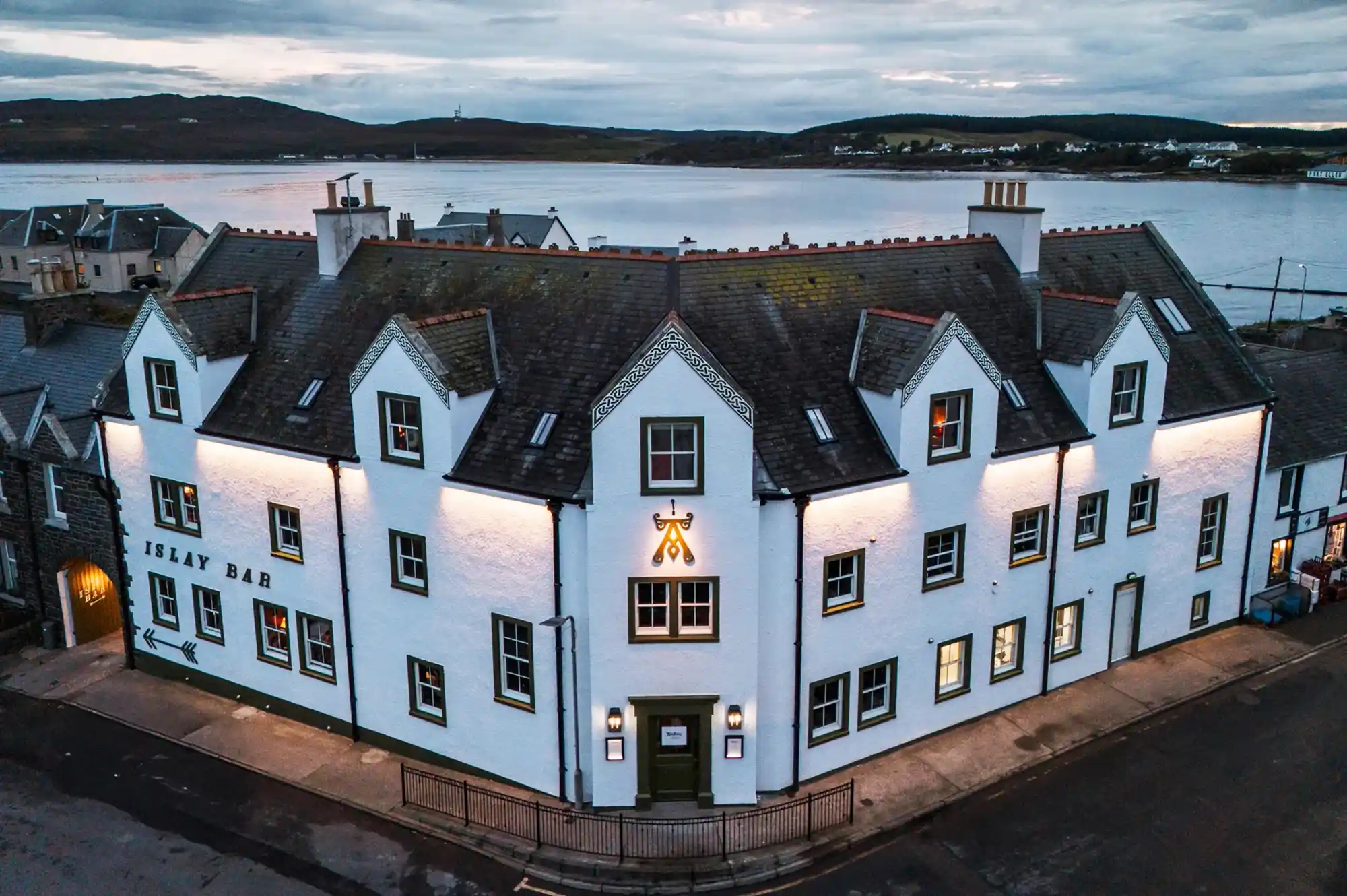 A white-walled building labeled Islay Bar with lit windows reflected in the calm waterfront at sunset.