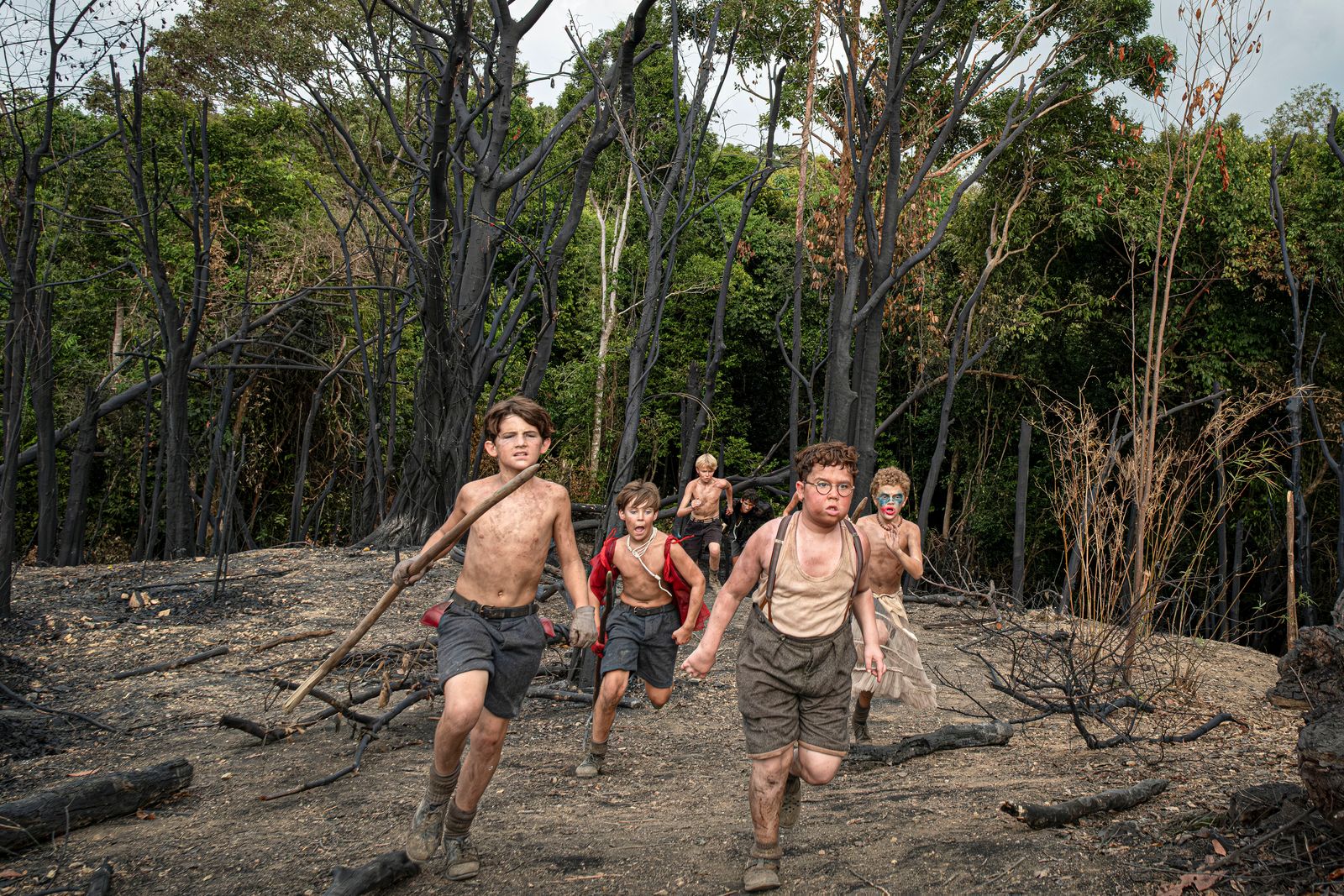 Group of young people holding makeshift spears and chasing through a natural landscape.